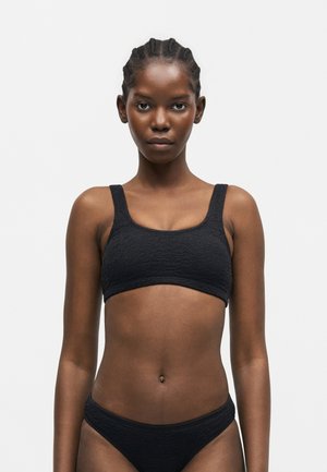 Jeune femme aux cheveux tressés portant un bikini noir texturé, debout devant un fond blanc uni, tournée vers l'avant.