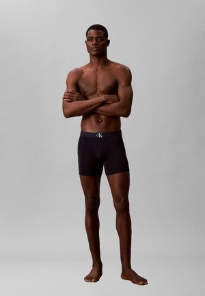 Athletischer Mann, barfuß stehend mit verschränkten Armen, trägt schwarze Calvin Klein Boxershorts vor einem schlichten grauen Hintergrund.