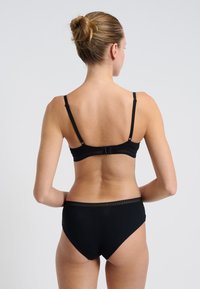 Woman with blonde hair in a bun wearing black bra and matching high-waisted underwear, standing against a plain light background.