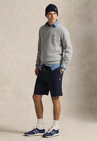 Homme portant un sweat-shirt gris, une chemise en denim bleu, un short bleu marine, un bonnet bleu marine, des chaussettes blanches et des baskets bleues, debout les mains dans les poches.