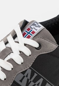 Sneaker grigia e nera con lacci bianchi, caratterizzata da una parte superiore testurizzata e un'etichetta con una bandiera norvegese e il logo. Materiali durevoli utilizzati.
