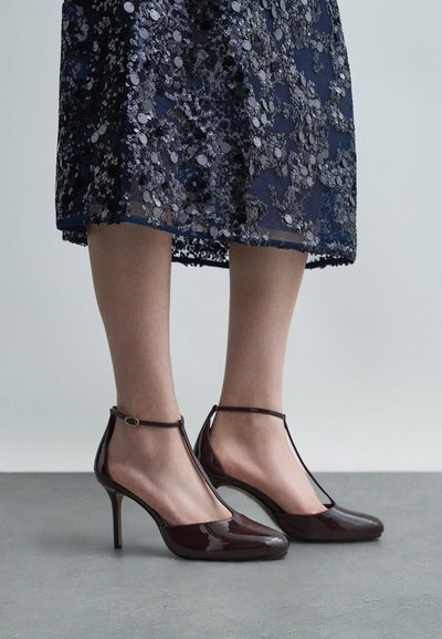 Καφέ παλτό δέρμα ψηλοτάκουνα παπούτσια με σχέδιο T-strap, που διαθέτουν μυτερή μύτη και λουράκι στον αστράγαλο με χρυσό τόνο κούμπωμα.