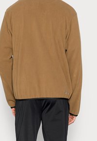 Brun fleecejacka med rund halsringning, långärmad och med svart kant vid fållen, med en diskret logotypmärke nära nederkanten på vänster sida.