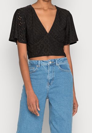 Frau trägt ein schwarzes, ausgestanztes, kurz geschnittenes Oberteil mit flatternden Ärmeln und hoch taillierten hellblauen Jeans aus Denim vor schlichtem Hintergrund.