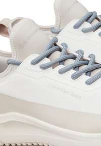 Zapatillas gruesas en blanco y beige con cordones grises y suelas de goma. Presentan la marca "Calvin Klein Jeans" en el lateral y un acabado texturizado.