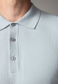 Světle modrá pletená polo košile s ribbed límcem, třemi šedými knoflíky a jemnou texturou. Detailní záběr na výstřih a légu knoflíků.