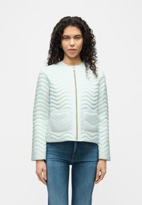 Γυναίκα με κατσαρά μαλλιά που φοράει ανοιχτό μπλε μπουφάν με τσέπες μπροστά και μπλε τζιν, στέκεται μπροστά σε απλό φόντο.