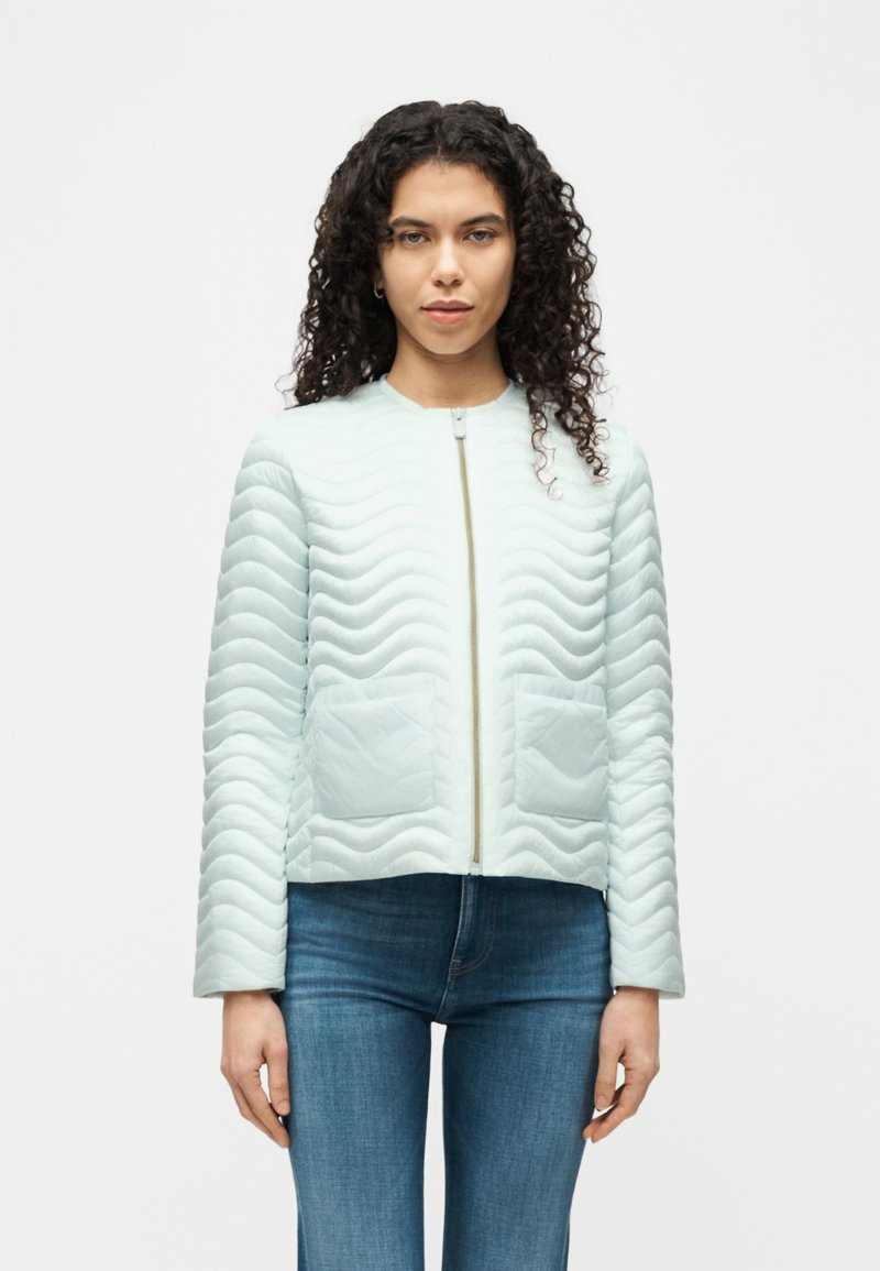 Γυναίκα με κατσαρά μαλλιά που φοράει ανοιχτό μπλε μπουφάν με τσέπες μπροστά και μπλε τζιν, στέκεται μπροστά σε απλό φόντο.