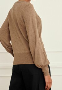 Pull en maille beige avec une surface texturée, doté de manches bouffantes et de poignets côtelés, associé à un pantalon noir.