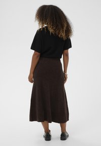 Črn kratki rokav z zgornjim delu, kombiniran z rjavim volnenim krilom do kolen, ki poudarja teksturirano površino in sproščen kroj. Črne čevlje dopolnjujejo celoten videz.