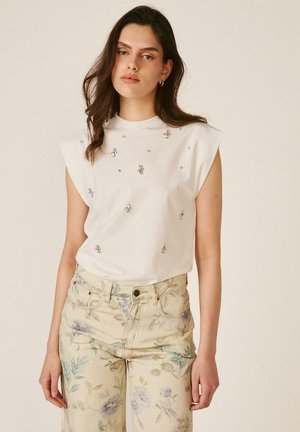 Mujer con cabello largo y oscuro, vestida con una blusa blanca sin mangas adornada con pequeñas gemas y pantalones de color claro con un patrón floral.