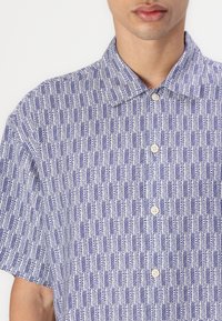Camisa de manga corta con un patrón geométrico en azul y blanco, diseño con cuello y cierres de botones, hecha de una tela texturizada.