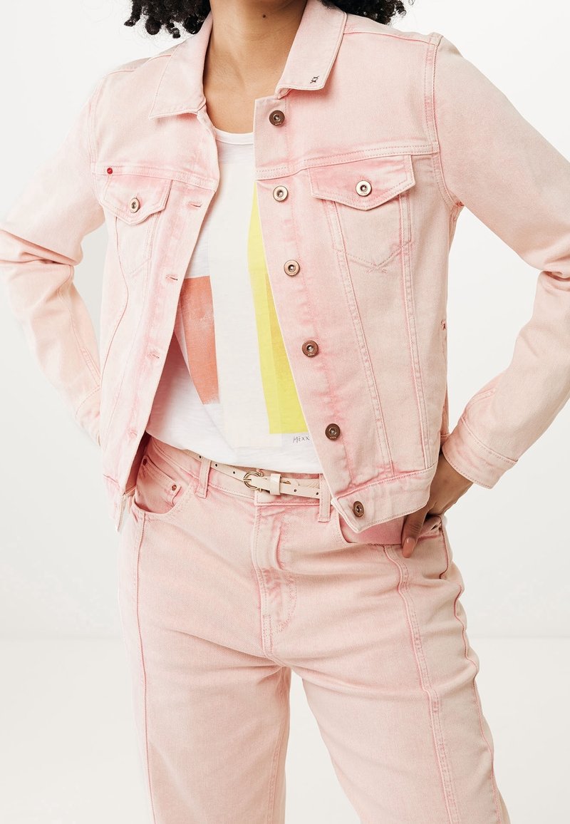 Personne portant une veste en jean rose clair et un pantalon avec un t-shirt blanc à motif graphique et une ceinture beige, debout les mains sur les hanches.
