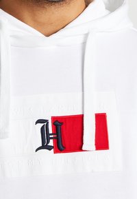 Gros plan d'un sweat à capuche blanc avec un logo rectangulaire rouge et blanc et une lettre H noire brodée sur la poitrine.