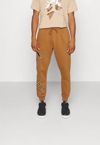 Man med bruna joggerbyxor med texten "JORDAN" på ena benet, beigefärgad Jordan-t-shirt och svarta Nike-sneakers mot en enfärgad bakgrund.