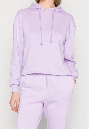 Woman wearing lavender hoodie with drawstrings and matching lavender sweatpants, one hand in pocket against plain background.