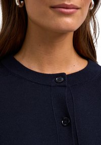 Marineblaue Strickjacke mit rundem Kragen, die zwei schwarze Knöpfe am Ausschnitt und eine strukturierte Oberfläche aufweist.