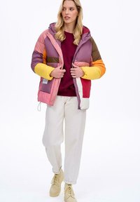Kolorowa kurtka puchowa z mieszanką paneli różowego, żółtego i brązowego. Z kapturem, zapinana na zamek, zestawiona z kremowymi spodniami i beżowymi botkami.