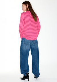 Lyserød, langærmet top med striber, kombineret med løse blå jeans og sorte ankelstøvler med hæl. Set bagfra med fokus på den afslappede pasform og kontrasterende farver.