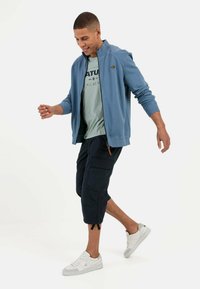 Lichtblauwe zip-up jas over een grijs T-shirt met zwarte tekst, gecombineerd met donkere cargoshorts en witte sneakers. Eenvoudig en casual ontwerp.