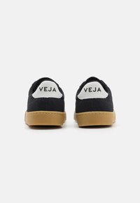 Tênis pretos de lona com sola de borracha, apresentando uma aba no calcanhar branca com o logótipo "VEJA". Design arredondado, tecido texturizado e estilo minimalista.