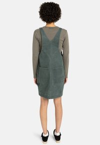 Abito da salopette in denim verde con schiena a V, dotato di due tasche frontali e orlo a lunghezza ginocchio. Indossato sopra una maglietta a maniche lunghe.