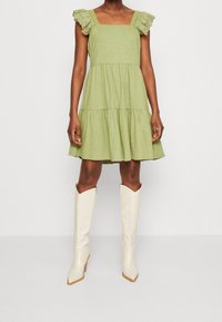 Groen gelaagde jurk met gerimpelde, met kant afgewerkte bandjes en een vierkante halslijn, gecombineerd met witte, kniehoge, puntige laarzen op een neutrale achtergrond.