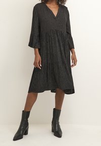 Robe noire avec un décolleté en V, des manches cloche et un design en couches. Présente des rayures métalliques et une coupe décontractée. Associée à des bottines.