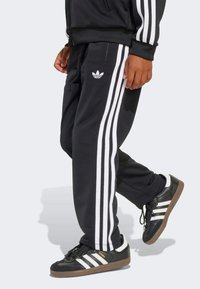Black athletic pants featuring three white stripes along the sides, with a small white logo on the front pocket, made from a smooth fabric.