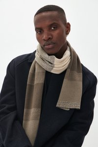 Un jeune homme portant un manteau noir et une écharpe en laine beige, avec une petite boucle d'oreille, regardant calmement l'objectif sur fond uni.