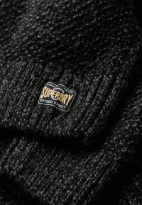 Zwarte gebreide beanie met een getextureerd patroon, voorzien van een gestikt label met "Superdry" in goud op een zwarte achtergrond.