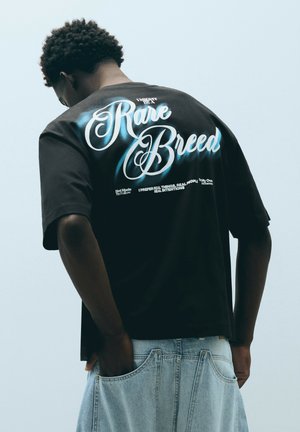 Persona che indossa una maglietta nera con "Rare Breed" in grande scrittura e jeans azzurri con le mani nelle tasche posteriori.