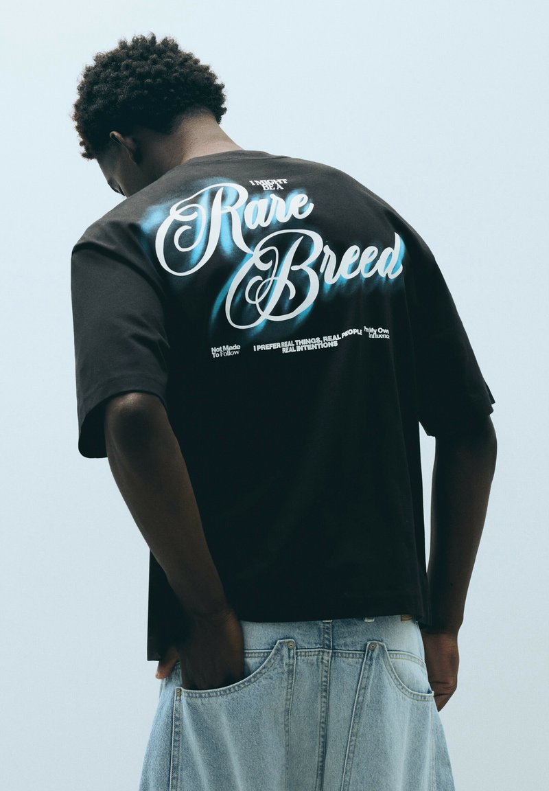 Person wearing a black T-shirt with "Rare Breed" in large script and light blue jeans with hands in back pockets.