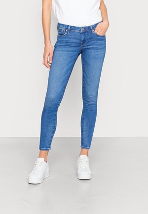 Persoon die blauwe skinny jeans en witte sneakers draagt, staat met één hand in de zak tegen een eenvoudige achtergrond.