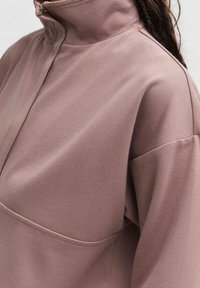 Sudadera de manga larga y cuello alto en tela suave color malva. Cuenta con un cuello con cremallera y un bolsillo frontal con un acabado liso y ligeramente texturizado.