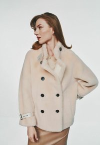 Dwurzędowy płaszcz z sztucznego futra w miękkim beżu z czarnymi guzikami, z szerokim kołnierzem i regulowanymi detalami na rękawach.