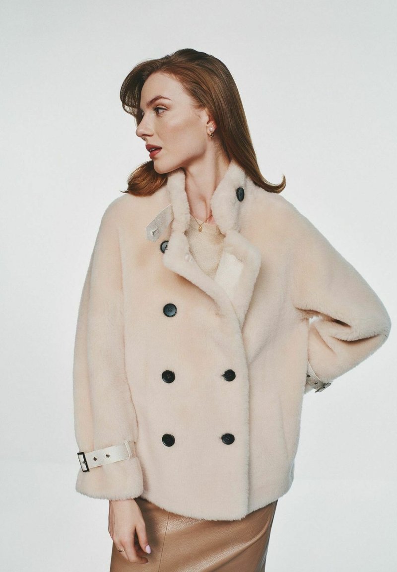 Dwurzędowy płaszcz z sztucznego futra w miękkim beżu z czarnymi guzikami, z szerokim kołnierzem i regulowanymi detalami na rękawach.