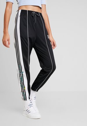 Zwarte Adidas trainingsbroek met witte zijstrepen, bloemenrankdetail op het been, gedragen met witte Adidas sneakers en witte sokken.