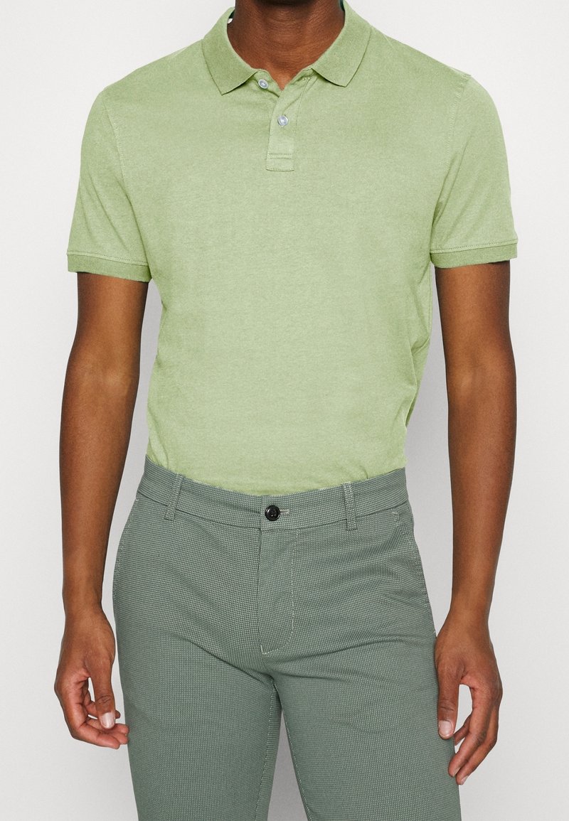 Homme portant un polo vert clair à manches courtes et un pantalon gris texturé, debout devant un fond uni.