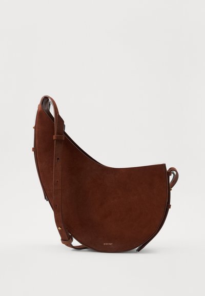Brun ruskinds skuldertaske med en halvmåneform, der har en slank justerbar rem og et minimalistisk design. Guld-branding detaljer synlige.
