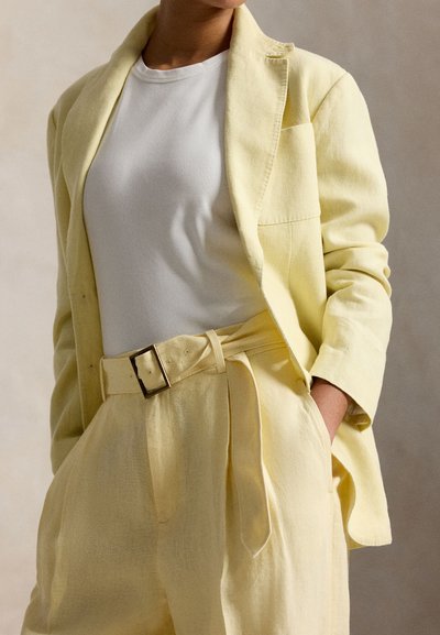 Άτομο που φοράει ανοιχτό κίτρινο παντελόνι με ζώνη και ταιριαστό σακάκι πάνω από λευκό ριμπ μπλουζάκι με στρογγυλή λαιμόκοψη, με το χέρι στην τσέπη.