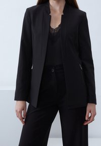 Zwarte op maat gemaakte blazer met een schone, open voorkant en zijzakken met rits. Gemaakt van een gladde stof, heeft een minimalistische silhouet.