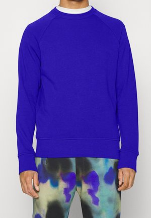 Mężczyzna w gładkiej królewsko-niebieskiej długiej bluzie i wielokolorowych spodniach tie-dye stoi na białym tle.
