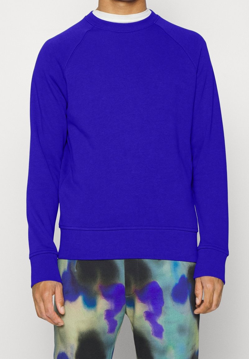 Homme portant un sweat-shirt uni bleu roi à manches longues et un pantalon tie-dye multicolore, debout devant un fond blanc.