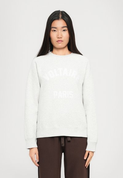 Ung kvinde med langt mørkt hår iført en lys grå sweatshirt med teksten "Voltaire Paris" og mørkebrune bukser, der står mod en ensfarvet baggrund.