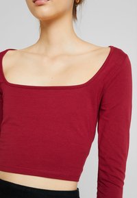 Burgundy pitkähihaine crop-top, jossa on neliönmuotoinen kaula-aukko, valmistettu pehmeästä kankaasta. Ominaisuuksiin kuuluu istuva malli ja sileä pinta.