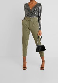 Schwarzes Pailletten-Oberteil mit langen Ärmeln und V-Ausschnitt, kombiniert mit olivgrünen, hoch taillierten Hosen mit einem Gürtel. Die Accessoires bestehen aus einer schwarzen Handtasche und hohen Schuhen.