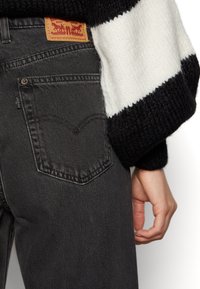 Jeans en denim noir avec une pièce en cuir marron arborant un logo, des coutures de poche arrière visibles et une manche en tricot texturé.