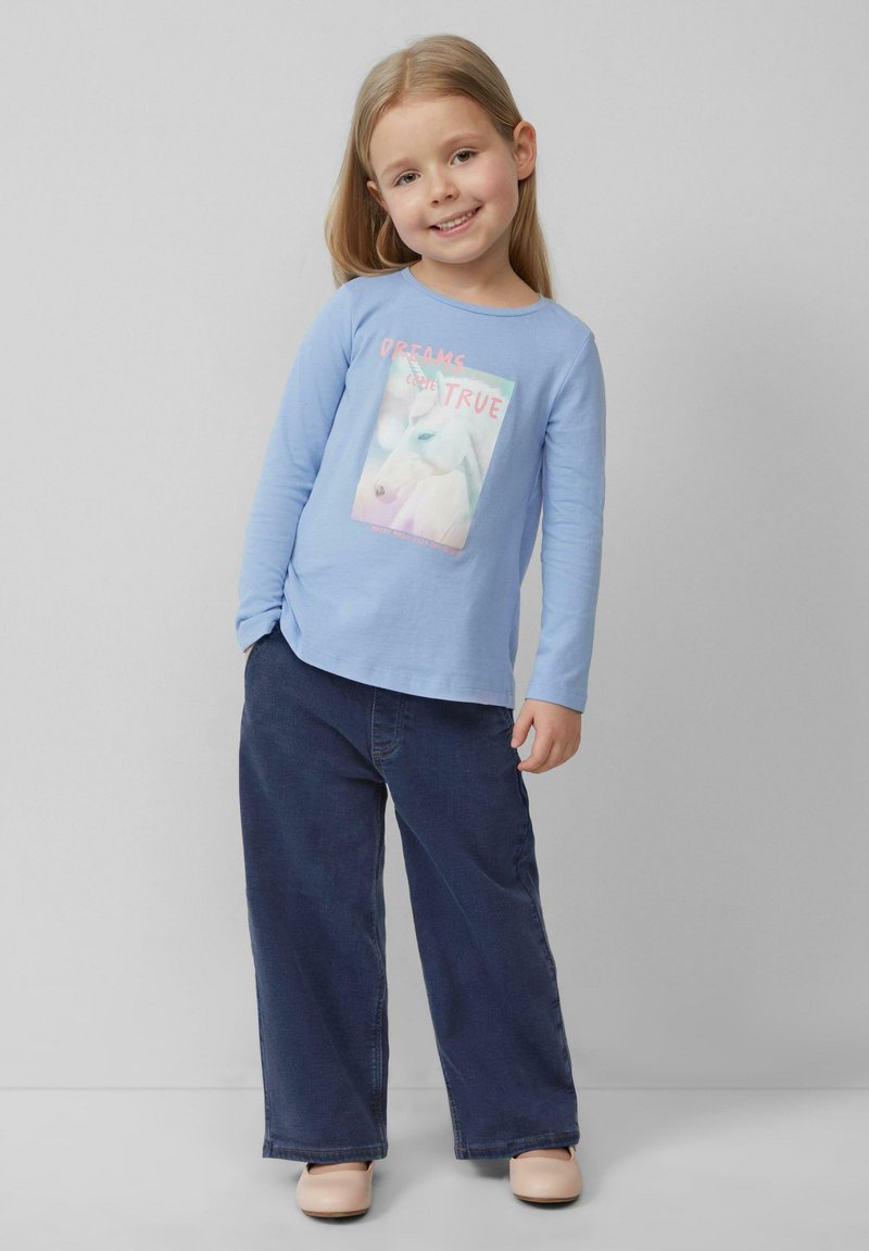 Lichtblauwe langwerpige blouse met een eenhoorn grafiek en de tekst "DREAMS COME TRUE", gecombineerd met donkerblauwe wijde jeans en beige schoenen.