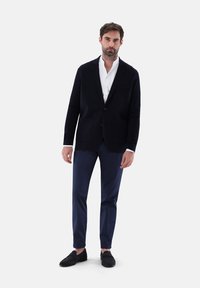 Marineblazer aus strukturiertem Stoff mit zwei Vorderaschen, kombiniert mit schmal geschnittenen marineblauen Hosen und schwarzen Loafers, über einem weißen Hemd.